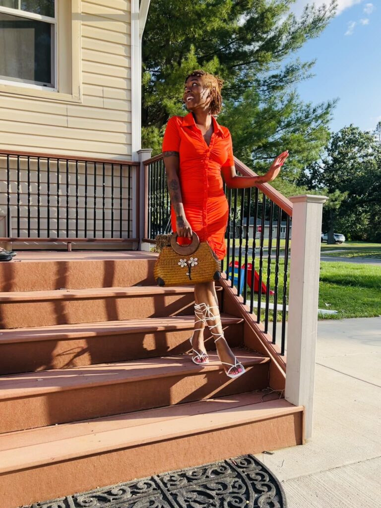 Elitrise posing on outdoor steps in a bright orange dress with white lace-up heels and a woven handbag.